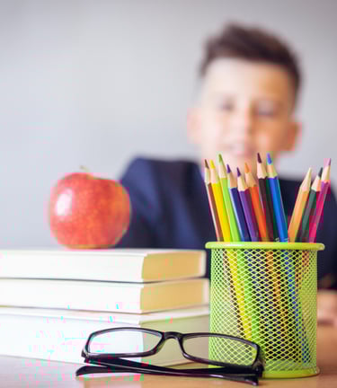 a boy sitting at a table with an apple, book, pencils and glasses