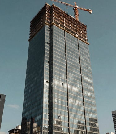 A low-angle high-resolution shot of a modern vertical glass building under construction with a crane visible, in a bustling Latin American / Spanish city, bright daylight, Muted Slate Blue sky, professional photography.
