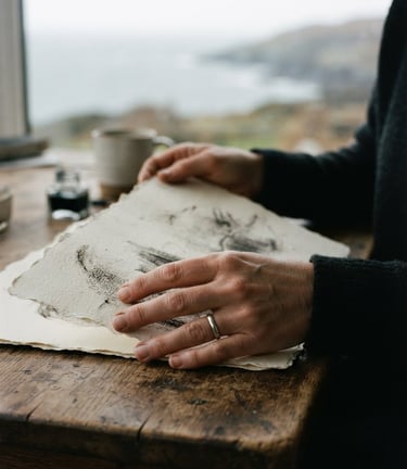 Close-up of Rachel's hands holding soft charcoal, textured watercolor paper on an oak table, diffused overcast coastal daylight