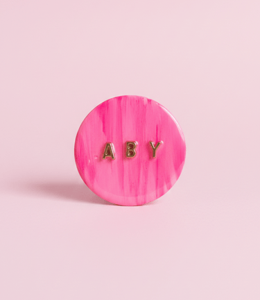 Pop socket para teléfono en tono rosa personalizado, con iniciales ABY en dorado.