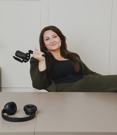 Adriana a podcaster in an olive blazer holding a microphone at her studio desk with headphones.