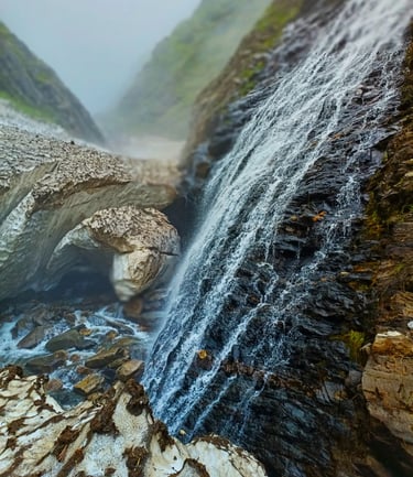 Snow-melt waterfall cascading over dark rocks in a misty mountain gorge