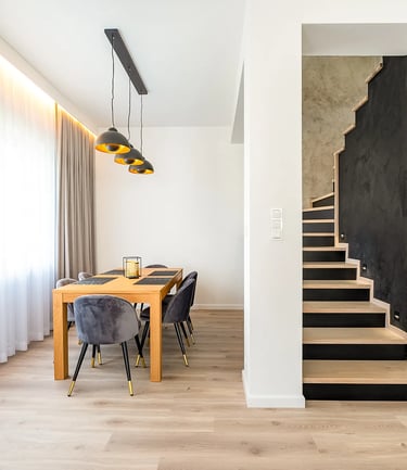 Jadalnia z drewnianym stołem i krzesłami, wiszące lampy, obok schody z czarną ścianą, mikrocement.