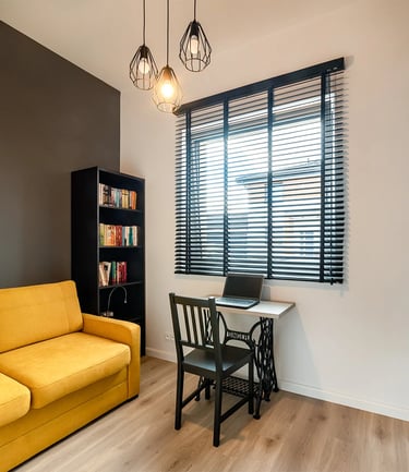 Pokój z żółtą sofą, biurkiem z laptopem przy oknie z żaluzjami i regałem.