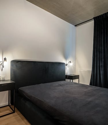 Minimalistyczna sypialnia z czarnym łóżkiem, dwiema lampkami nocnymi i zasłonami.