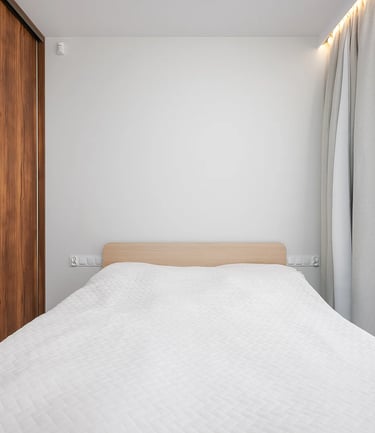 Łóżko z białą narzutą, drewniana szafa po lewej, okno z zasłonami po prawej.