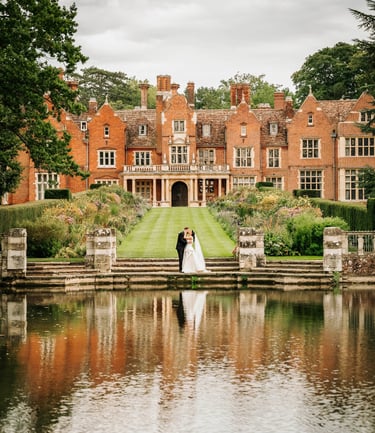 Bride and groom standing by a lake in front of a grand manor house