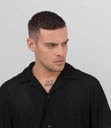 Portrait d’un modèle masculin en chemise noire sur fond gris clair, regard tourné sur le côté, ambiance studio épurée.