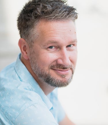 Portrait d’un comédien souriant en chemise claire, posé en extérieur avec un fond flou, idéal pour un book professionnel.