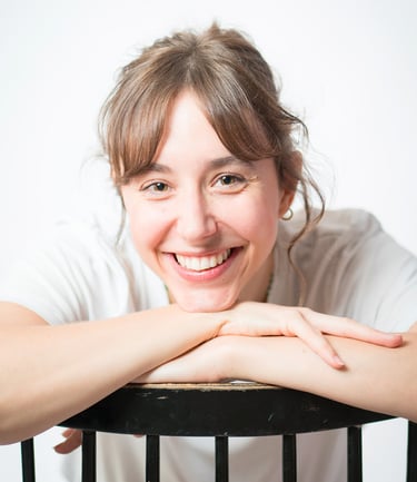 Portrait d’une comédienne souriante en studio, en t-shirt blanc, appuyée sur un dossier de chaise noire, sur fond blanc lumin
