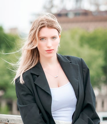 Portrait d’une modèle en veste noire, posant en extérieur avec un regard intense, sur fond flou de verdure et d’architecture 