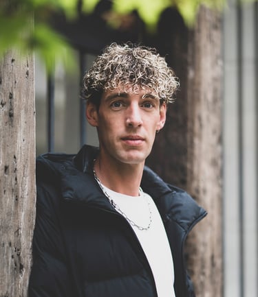 Portrait d’un modèle au regard intense, posé près d’un mur en bois, avec une lumière naturelle et un fond de feuillage flou.