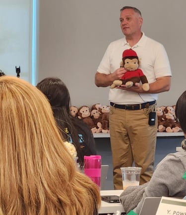 A man in a white polo shirt presents to a group while holding a Curious George plush toy.