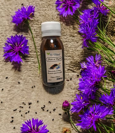 A bottle of Huile de Nigelle black seed oil surrounded by purple cornflowers and organic seeds.