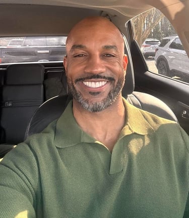 Smiling bald man with a salt and pepper beard wearing a green polo shirt inside a car.
