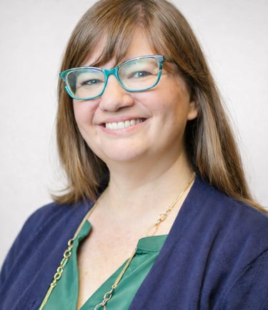 A smiling woman with brown hair and turquoise glasses wearing a navy cardigan and gold necklace.