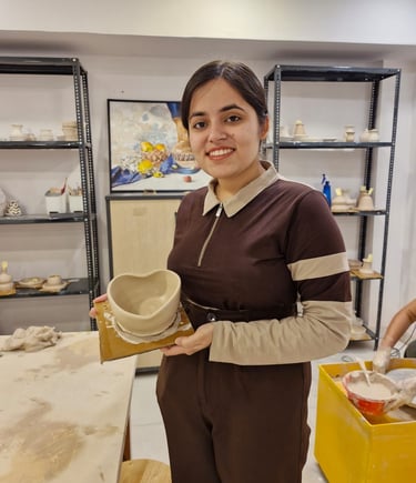 A smiling young woman holding her handmade clay bowl in a pottery classes studio with shelving.