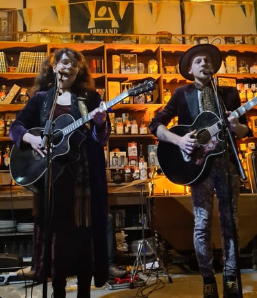 A folk music duo performing live with acoustic guitars in a rustic pub setting with warm lighting.