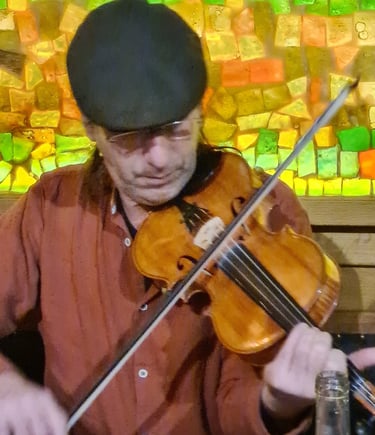 A musician playing a wooden fiddle in front of a colorful mosaic background at a live music pub.