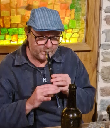 A man wearing a flat cap and glasses plays a traditional Irish tin whistle in a pub.