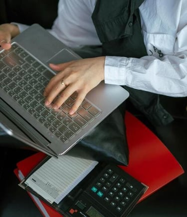 Una persona escribe en una laptop plateada junto a una calculadora y una carpeta roja