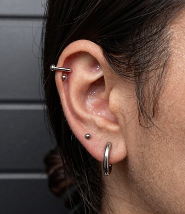 Close-up macro studio photography of a stylish Latin American / Spanish client in a high-end piercing studio, wearing polished implant-grade titanium ear jewelry and sleek silver studs against a dark charcoal metallic wall background with soft directional light.