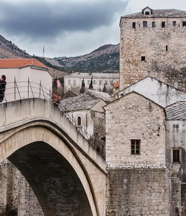 Tourists crossing Stari Most stone bridge in Mostar with mountains and cloudy sky