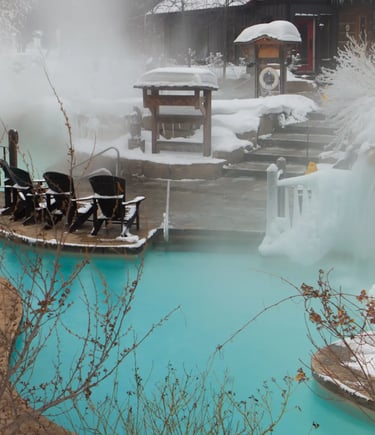 A large outdoor thermal spa complex with steaming pools surrounded by snow-covered mountains and pin