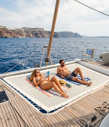 Couple sunbathing on catamaran bow netting with Greek island cliffs in background