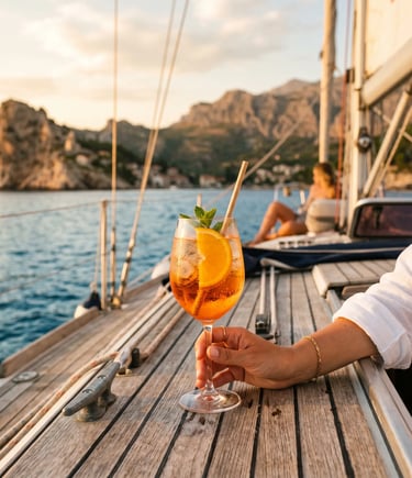 Hand holding Aperol Spritz glass on sailboat deck with rocky coastline at golden hour