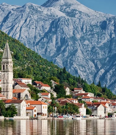 Stone village of Perast reflected in calm bay water with snow-capped mountains behind