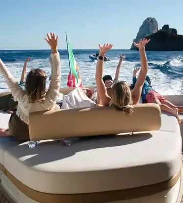Guests with arms raised on yacht aft deck approaching Faraglioni rocks off Capri