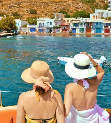Two women in sun hats on catamaran approaching colorful Klima fishing village in Milos