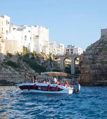 Small private boat with tourists exploring dramatic Polignano a Mare cliffs, Puglia coast.
