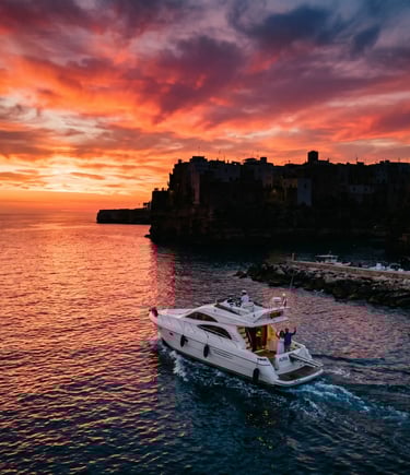 White private yacht leaving Polignano a Mare harbor under dramatic red sunset sky