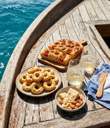 Taralli, focaccia barese, cheese, almonds and prosecco on wooden boat deck over turquoise sea.