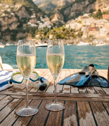 Two Prosecco glasses on teak boat deck with snorkeling gear and Positano coastline behind