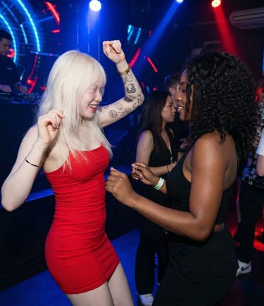 Two diverse women dancing at a lively nightclub with neon lights and a DJ performing in the background.