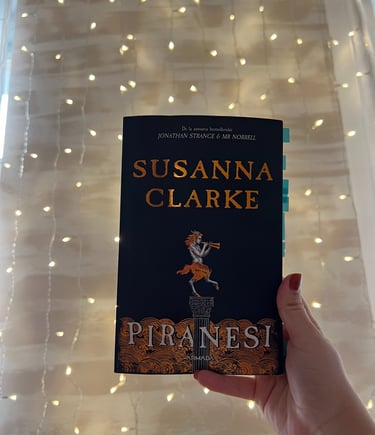 Mână ținând cartea 'Piranesi' de Susanna Clarke pe un fundal de luminițe de basm.
