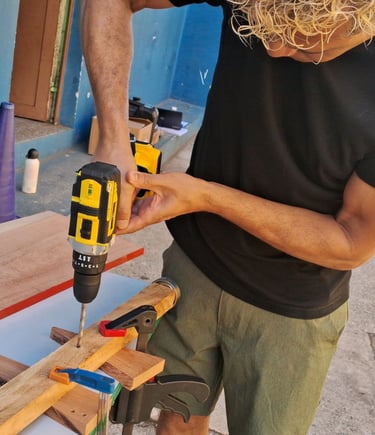 A woodworker uses a cordless electric power drill to bore holes in a wooden plank secured with clamps.