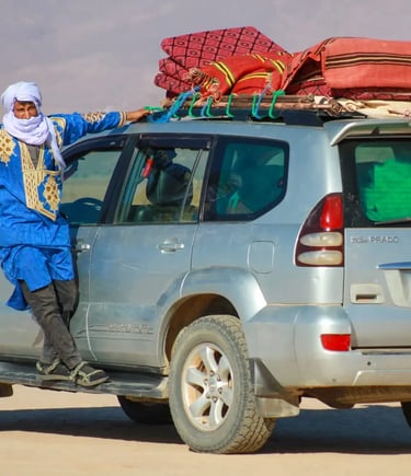 A man in traditional nomad dress stands on the running board of a 4x4 during a Moroccan Sahara trek
