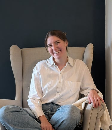 A smiling woman in a white linen shirt and jeans sits cross-legged in a beige wingback chair.