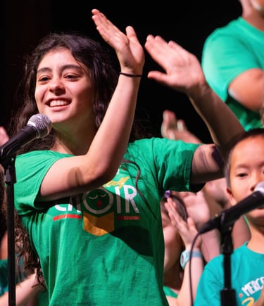 A smiling young girl in a green shirt claps during a live children's choir performance on stage.