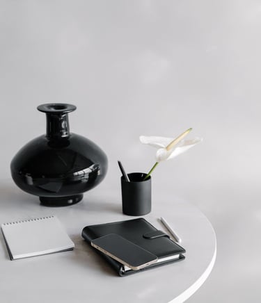A black vase, pencil cup with a a pen and flower in it, and notebooks on a table