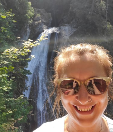Smiling woman with sunglasses taking a selfie in front of a scenic mountain waterfall.
