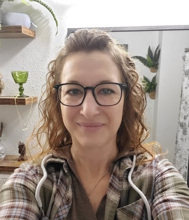 Woman with curly hair and black glasses wearing a plaid flannel shirt in a room with plants.