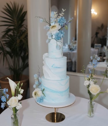 Three-tier white and blue marbled wedding cake decorated with fresh blue and white flowers on a gold stand.