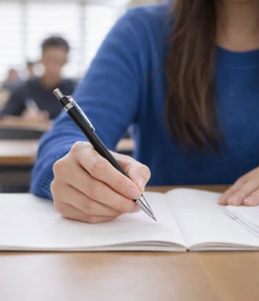A student in a blue sweater writes notes in a notebook with a pen during a university lecture.