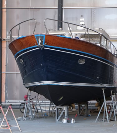 A classic wooden motor yacht with a blue hull undergoes maintenance and painting in a dry dock shipyard.