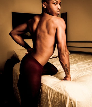 dark skinned young man shirtless in tight pants leaning against bed looking seductively at camera